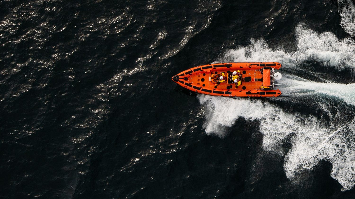 Aerial view of a lifeboat marked “B-016” speeding across dark, choppy water, leaving a white foamy wake behind. Several crew members in helmets and protective gear are visible on board as the vessel travels from left to right.