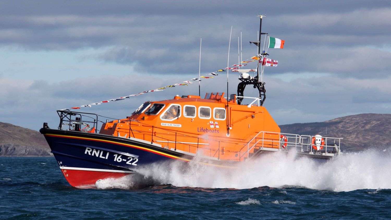 Lifeboat Alan Massey in choppy water, creating a large spray as it moves. Cloudy skies and distant hills appear in the background.