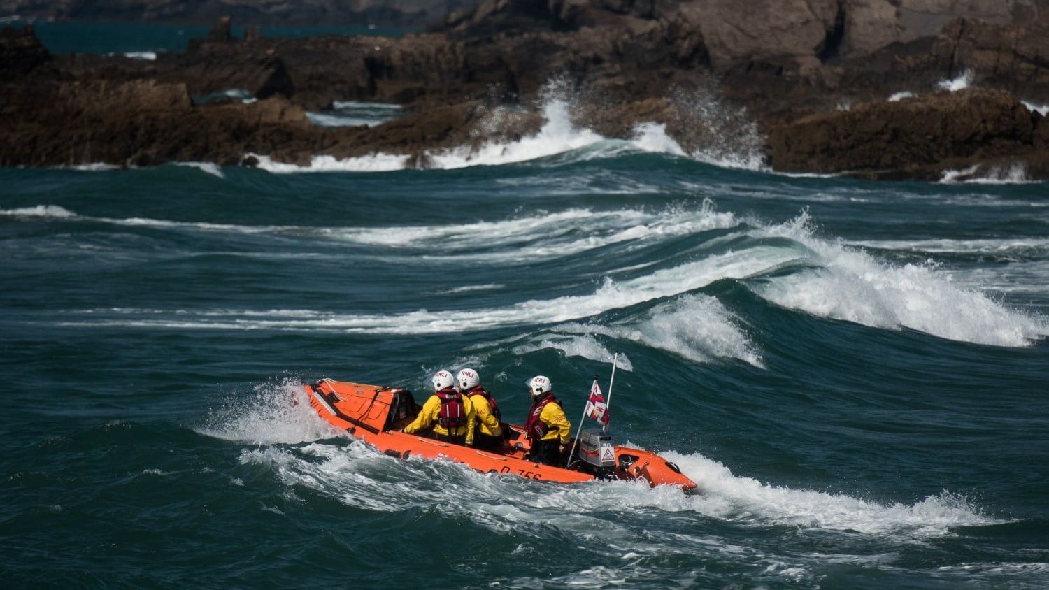A small rib with three crew in large blue waves with a rocky outcrop behind them.