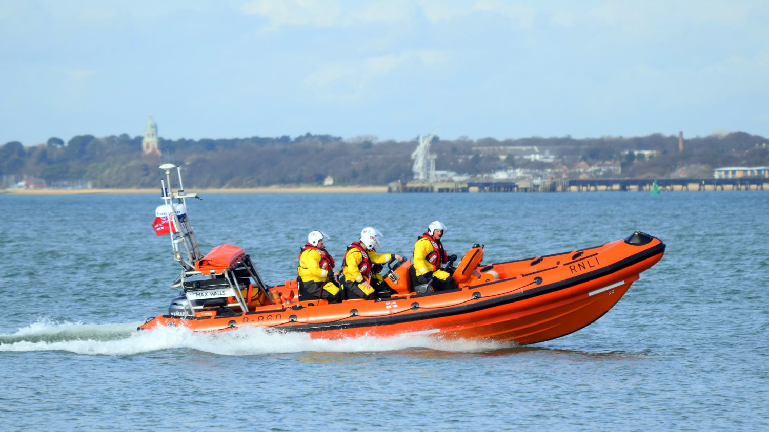 An B class lifeboat with four crew members moving across calm water. A coastline and buildings are visible in the background under a bright sky.