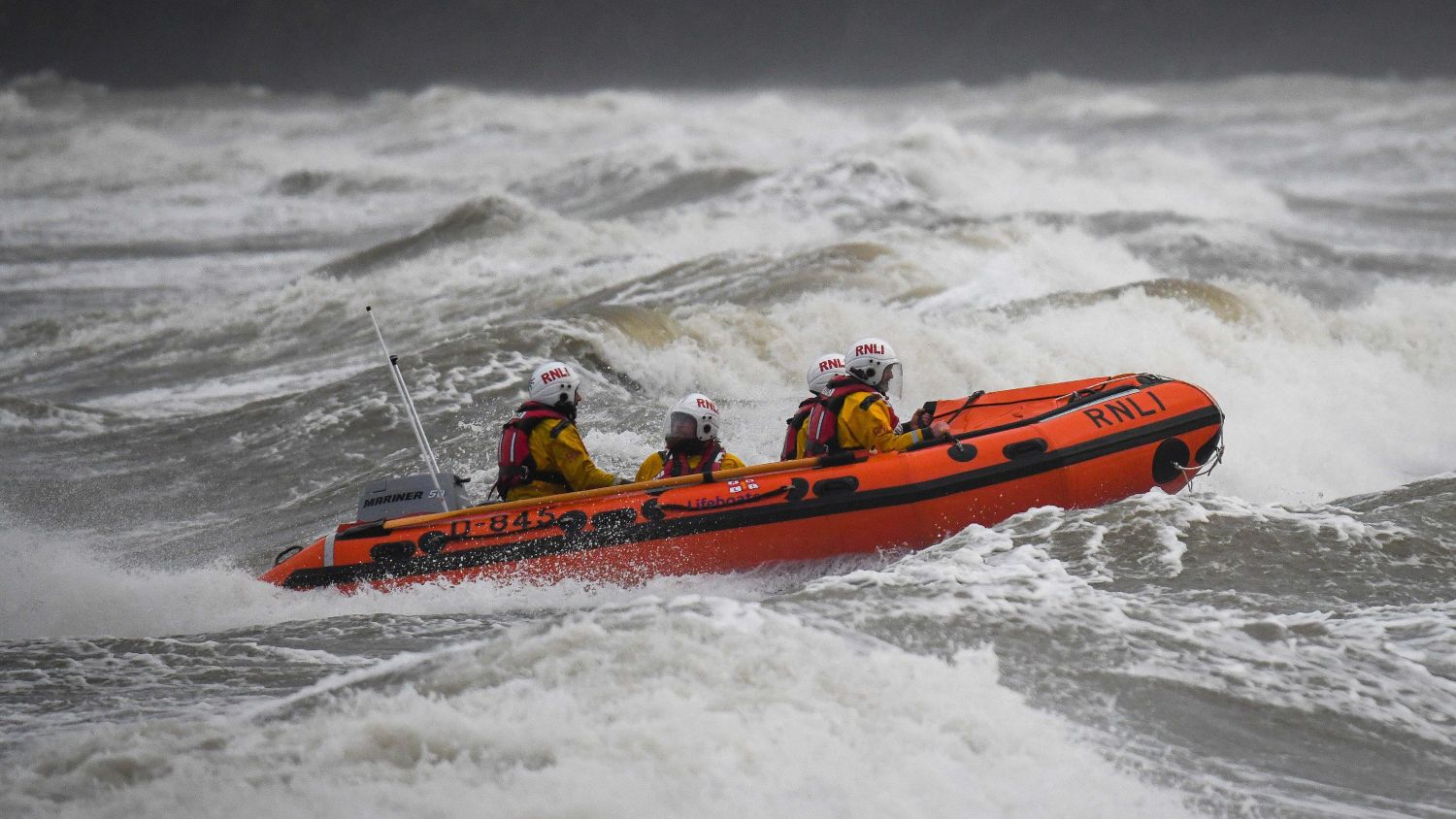 A D class lifeboat with four crew members moves through rough, breaking waves under a dark sky.