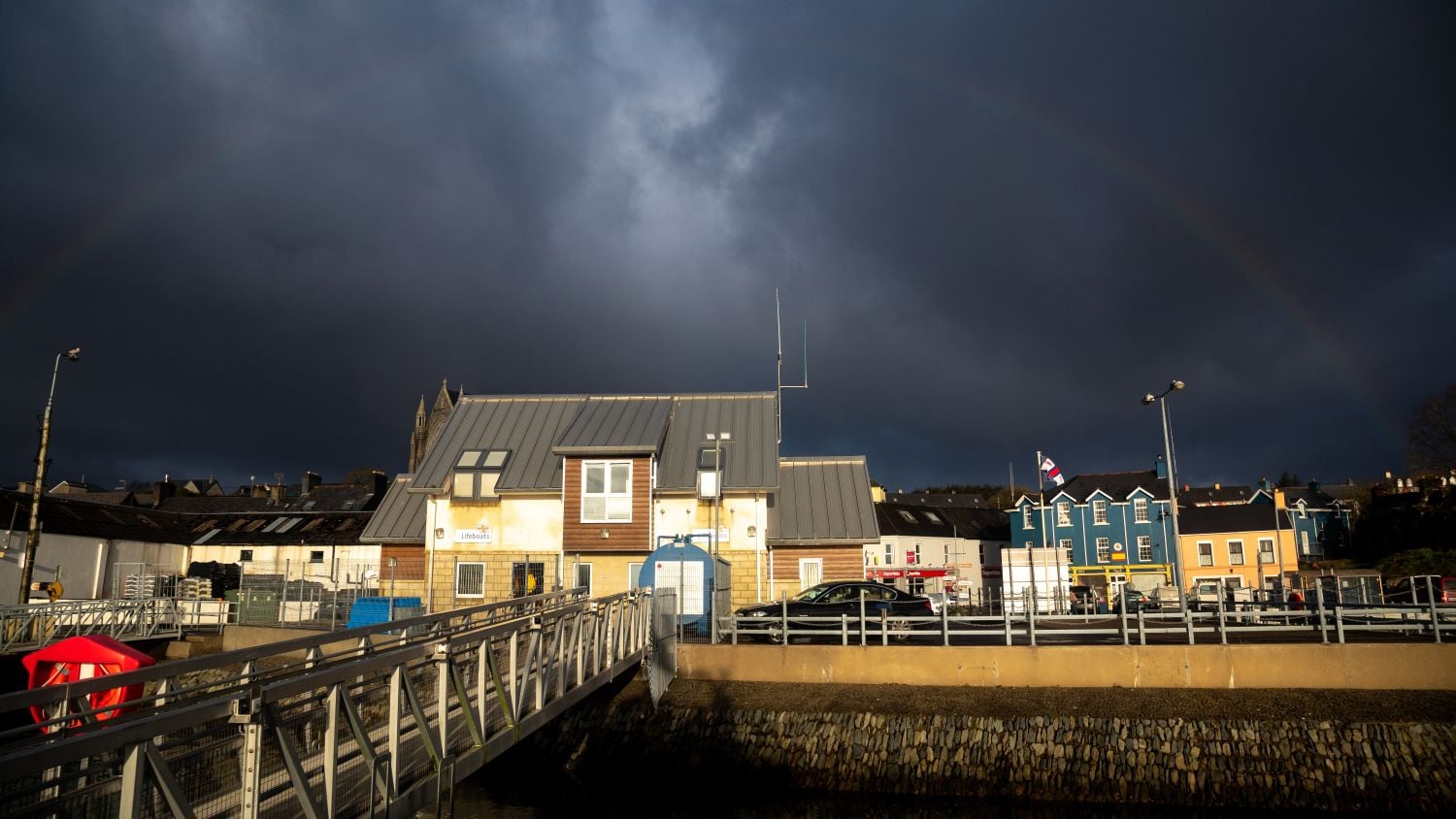 Castletownbere Lifeboat Station under dark clouds, with a faint rainbow overhead. In the background there are colourful waterfront houses.