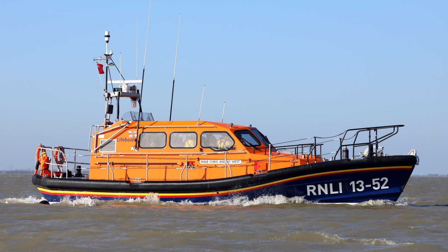 An orange RNLI lifeboat with the identification number RNLI 13-52 in across choppy brown water under a clear blue sky. The boat features a raised cabin with windows, railings along the deck, and rescue equipment visible at the rear.