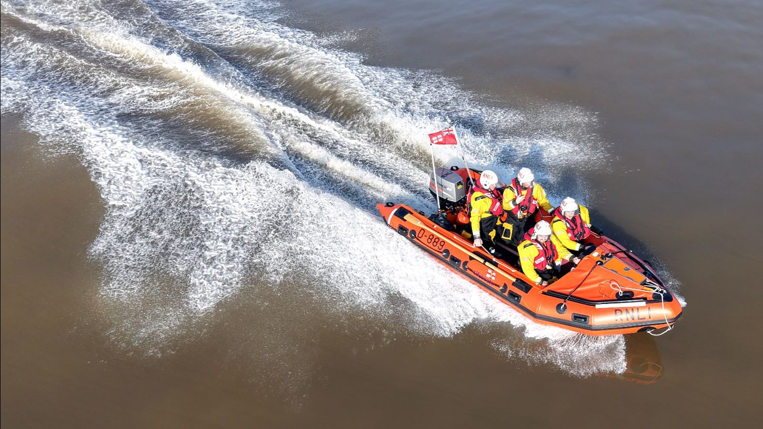 An aerial view of an orange RNLI lifeboat speeding across brown water, leaving a white wake as the crew sits secured on board.