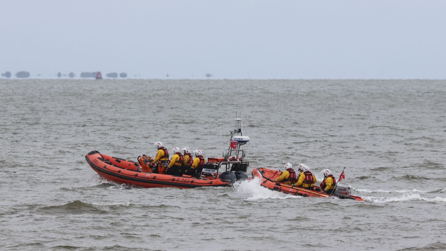 Two orange RNLI rigid inflatable lifeboats travel side by side across choppy grey sea, carrying crews in protective gear. The larger boat has it's name "Loving You" visible on the side