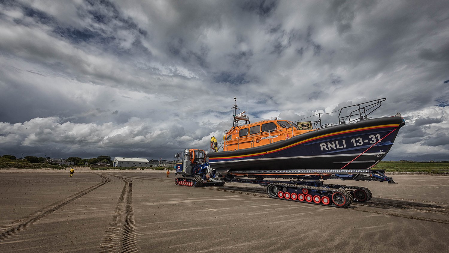 An RNLI Shannon‑class lifeboat (number RNLI 13‑31) sits on a tracked launching rig on a wide, flat beach. The sand is wet and reflective, showing the deep tracks left by the vehicle.  The sky is cloudy with patches of blue, and the sea is calm in the distance.