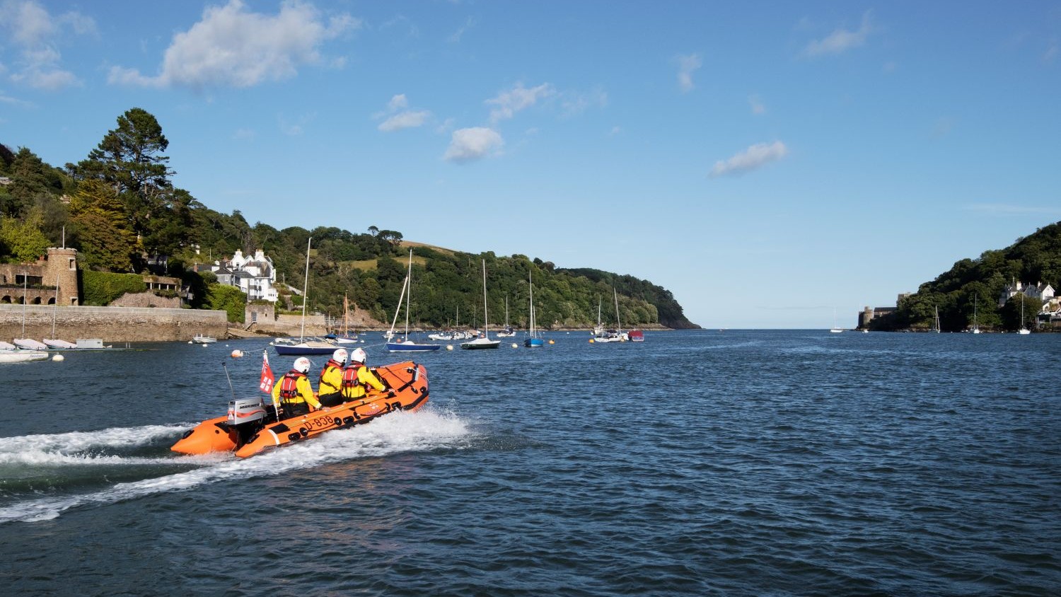 A small rib with three crew passes over calm blue water with forested hills either side of the water.