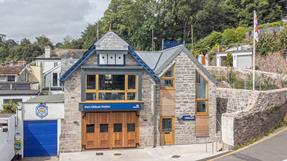 A two-storey stone building with large windows and wooden boathouse doors.