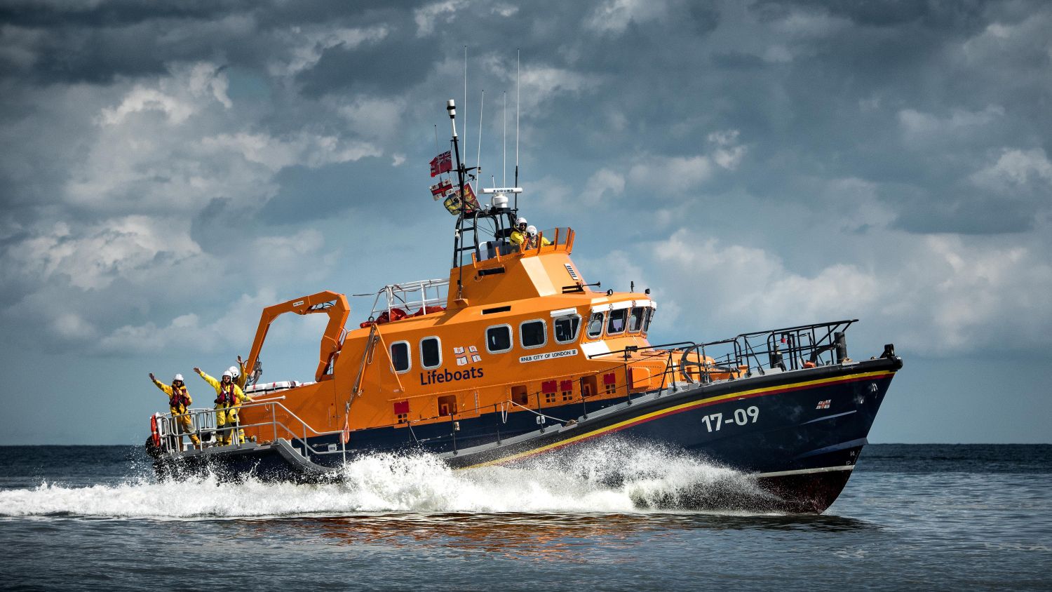 A lifeboat travelling across the water at speed, with several crew members visible on deck and dark clouds in the sky.