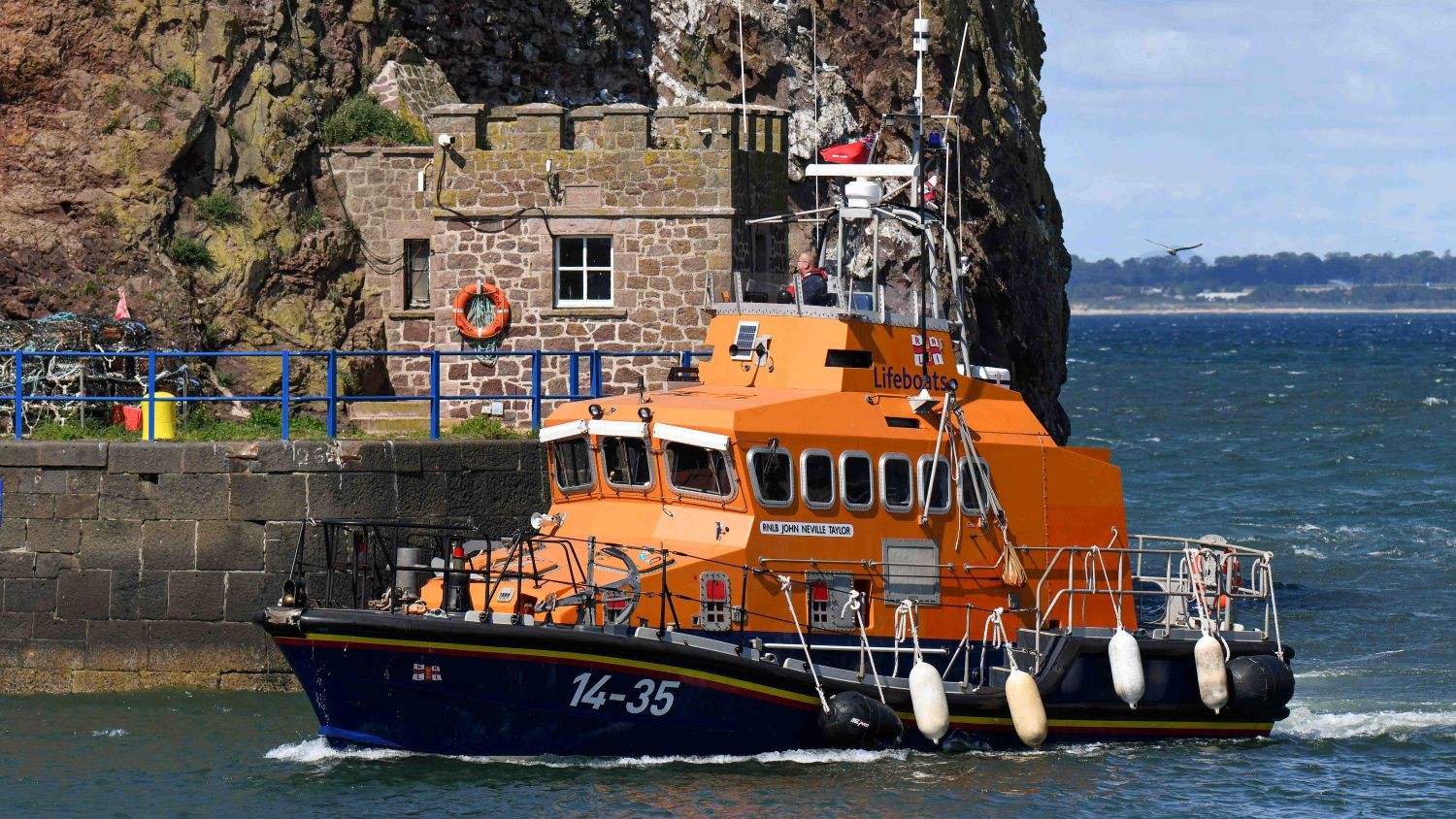 A Trent class all weather lifeboat passing a rocky cliff and sea wall.