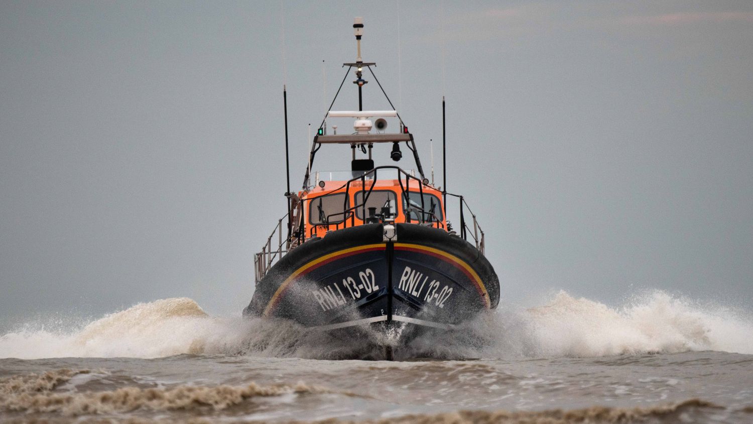 A lifeboat labelled ‘RNLI 13‑02’ is moving directly toward the camera through uneven water, creating visible spray on both sides. The sky is overcast.