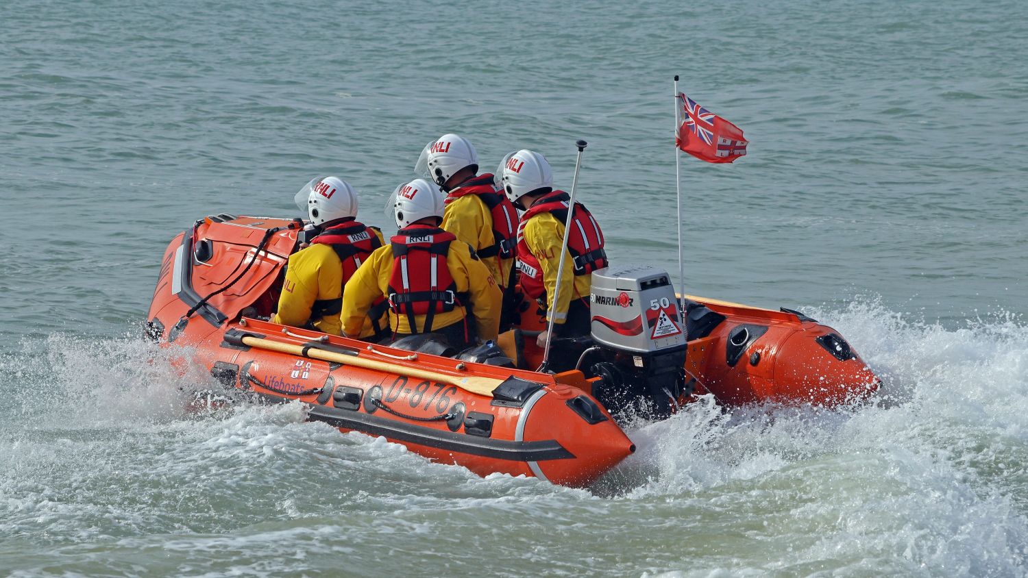 An inshore lifeboat with multiple crew members in protective gear moves through the water, creating a small wake behind it.