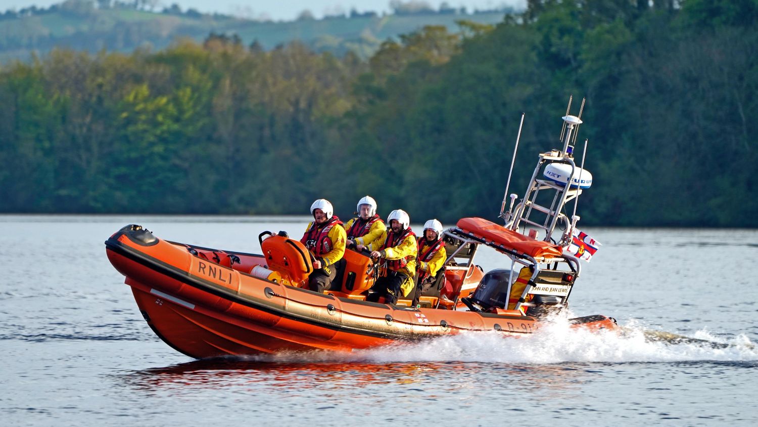An orange rigid inflatable lifeboat in calm water creating a small wake. Trees line the shoreline in the background, with hills visible beyond.