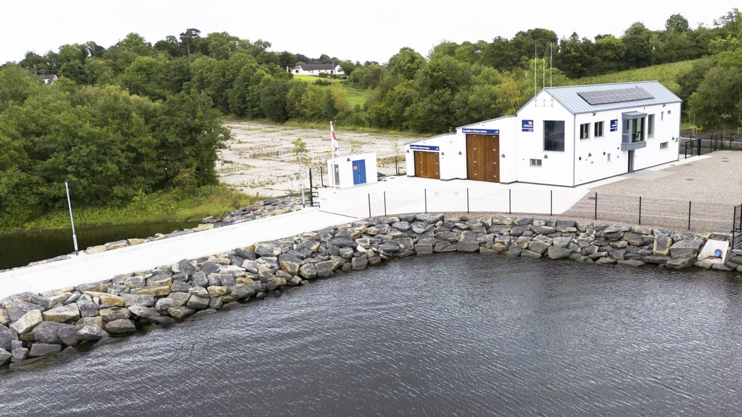 A white building with two boat sheds and a rock sea wall extending into the water.