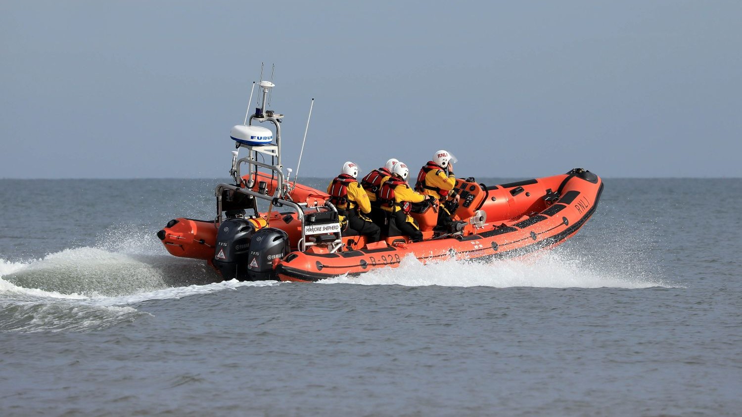 An orange RNLI inshore lifeboat speeds across calm open water, creating a white spray behind it. Four crew members, all wearing yellow waterproof jackets, red lifejackets, and white helmets, are seated. The sea is grey‑blue and the horizon is clear beneath a pale sky.