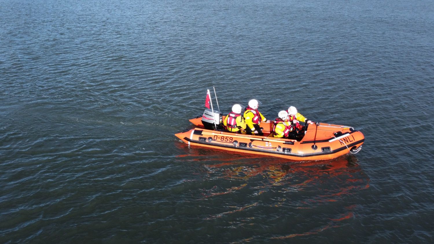 An orange RNLI rigid inflatable lifeboat with four helmeted crew members in high-visibility jackets travels across calm open water, viewed from above, with identification markings visible on the side of the boat.