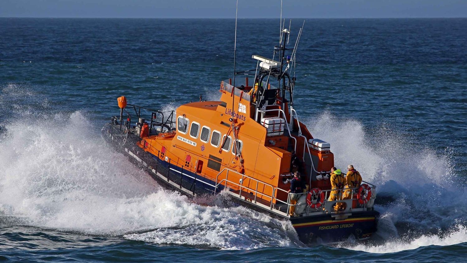 An Trent class all weather lifeboat creates spray on a blue sea.