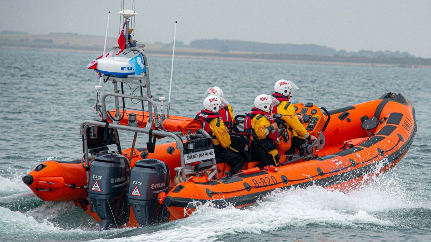 An RNLI inshore lifeboat travels at speed across choppy water, creating a white spray behind it. Four crew members wearing yellow waterproof jackets, red lifejackets, and white protective helmets are seated and holding onto the vessel’s framework. The lifeboat has two outboard engines at the rear, raised antennas, and a small radar unit mounted on a metal frame. In the background, a low, hazy coastline stretches across the horizon under an overcast grey sky.