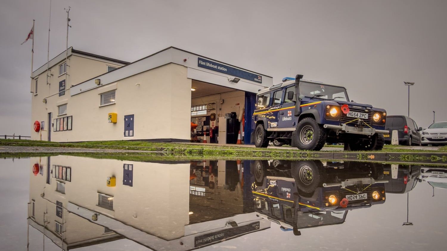 A one storey cream boatshed in front of a two storey cream building reflected in water on the ground.