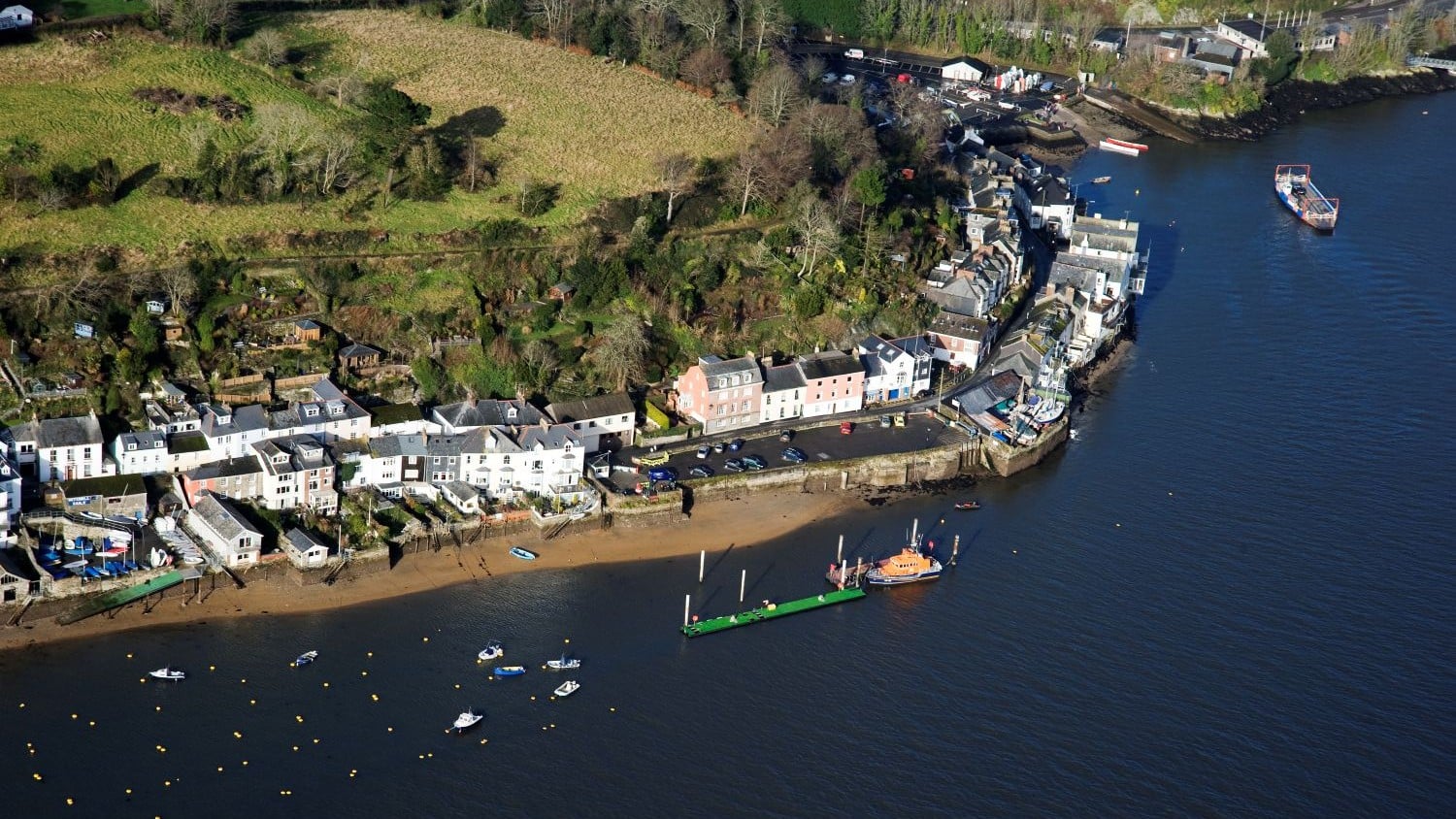 A road of houses along the waters edge with fields behind. An all weather lifeboat is moored afloat.