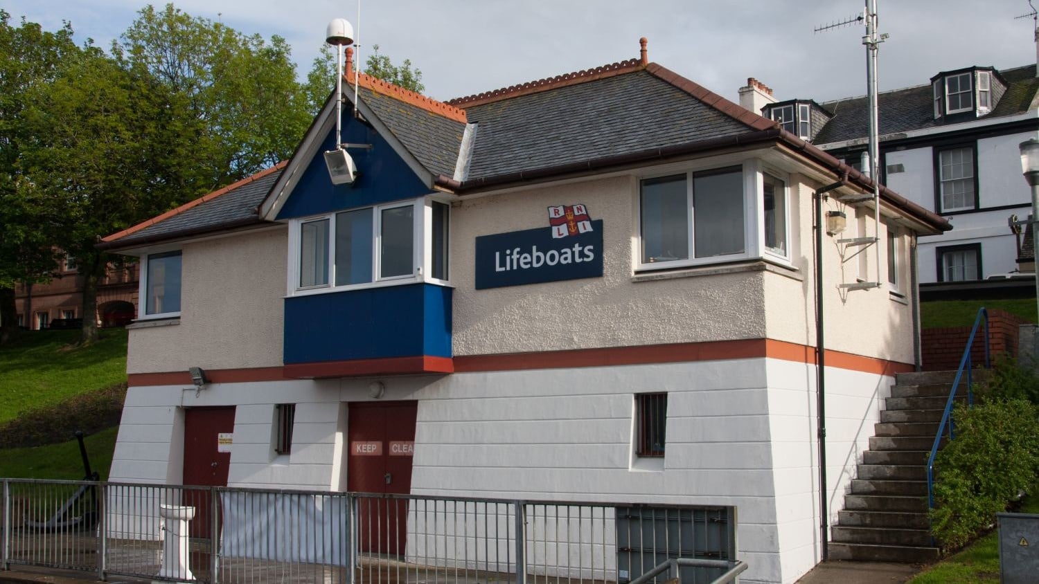 A two storey white, cream and blue building on a sea wall with steps running up a grassy bank behind it.