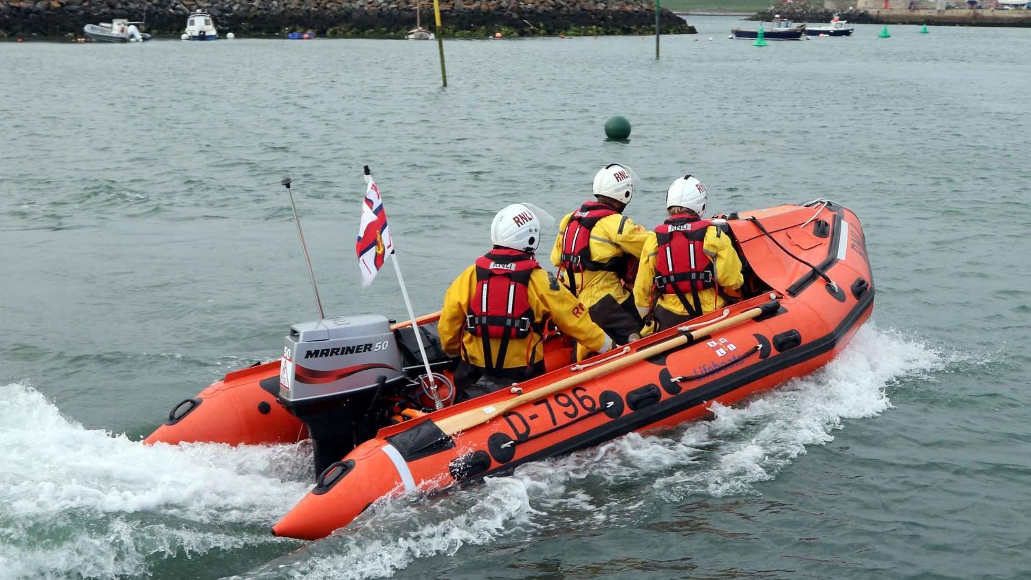 A small rib with three crew heading along a rocky sea wall.