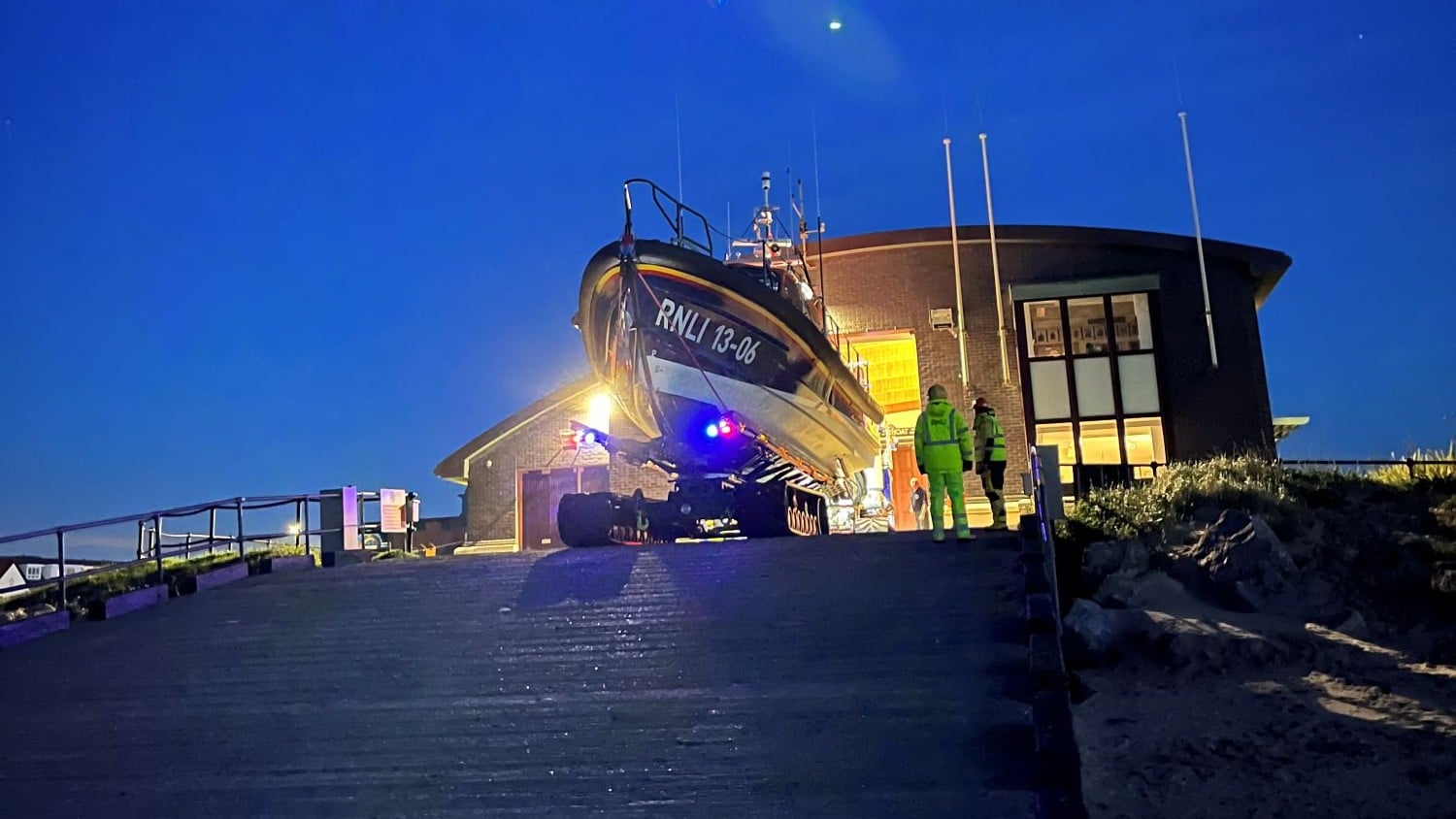 An All Weather Lifeboat at the top of a slipway with a two storey building with a curved roof behind it at dusk.