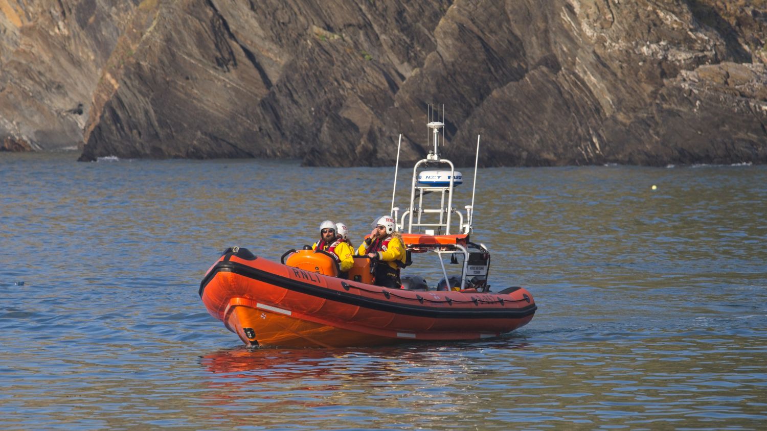 RNLI B class lifeboat  Patrick and Dianne O'Connor on calm coastal waters beneath steep rocky cliffs, with two crew members in full rescue kit on board and the orange hull reflected in the sea.