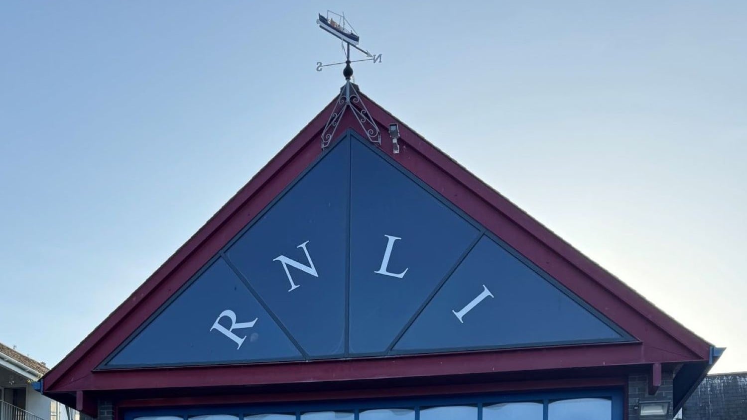 A blue triangular roof end with red edging and letters spelling out RNLI. There is a lifeboat weather vane on top.