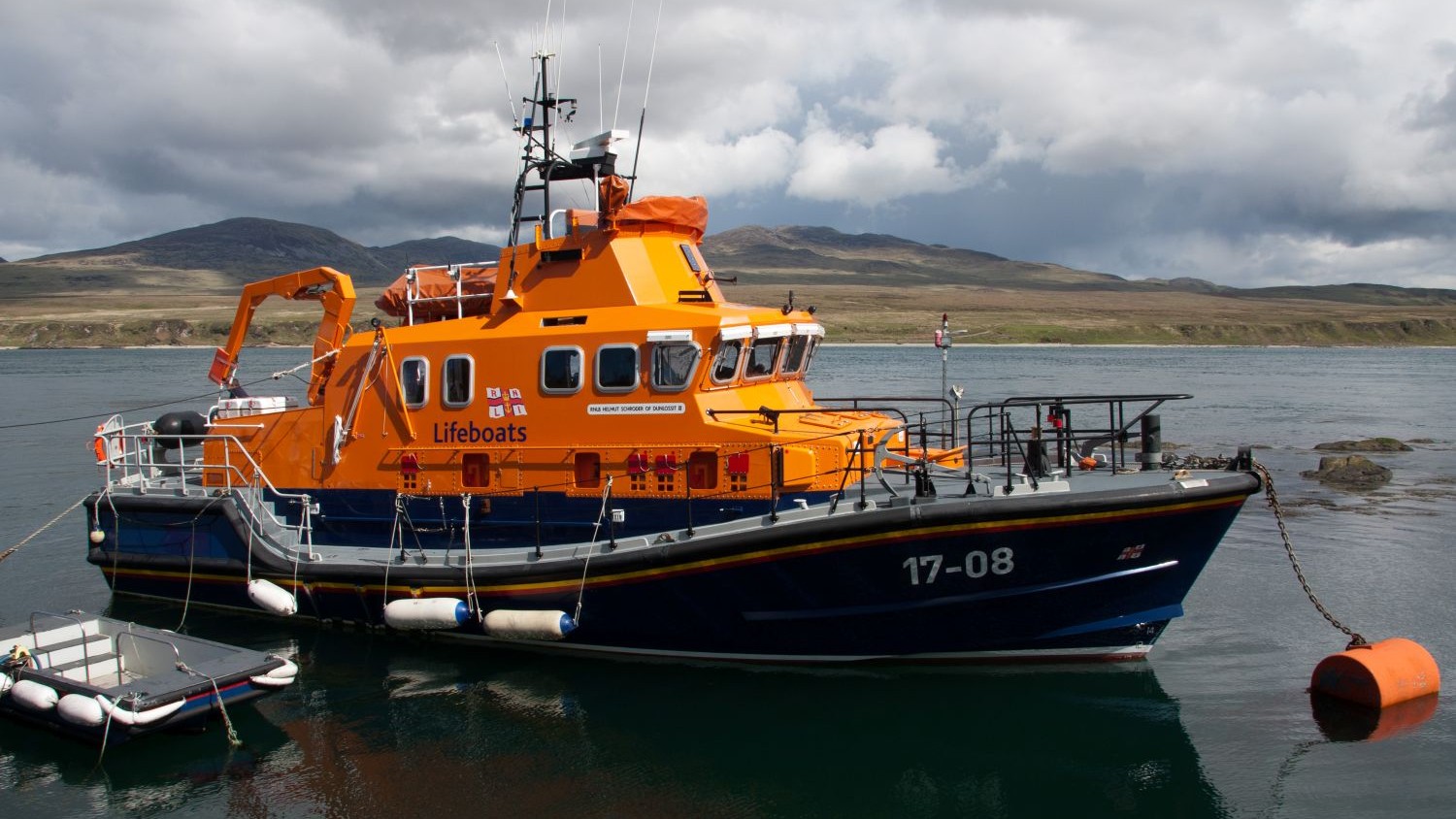 A lifeboat is moored in calm water near a rugged coastline. The boat has the identification number 17‑08 on the bow and a small inflatable dinghy floats next to the lifeboat. In the background, low hills and open land stretch beneath a cloudy, dramatic sky.