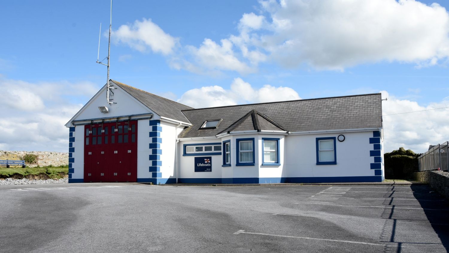 A one storey white building with blue edges and grey roof with large red doors to the boat shed.