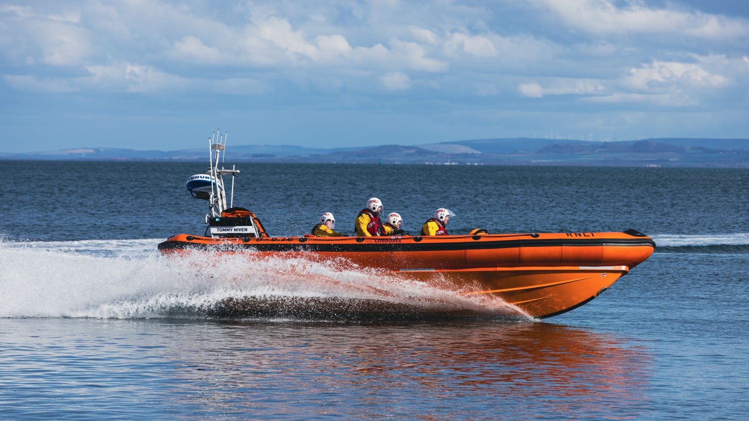 An orange rigid inflatable boat speeds across calm water, creating white spray, with four helmeted crew members in high‑visibility jackets seated onboar