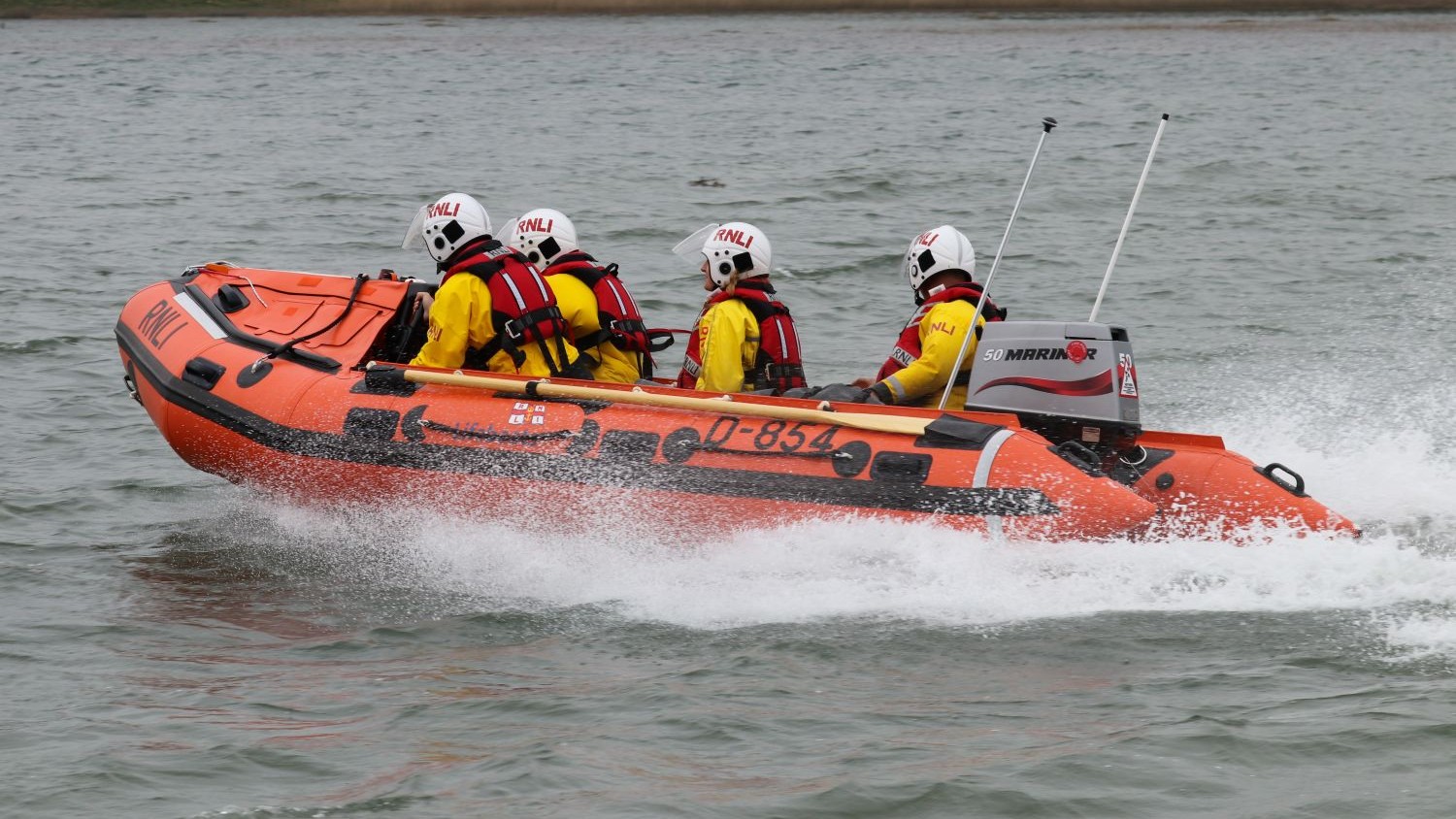 An orange RNLI inflatable lifeboat travels across the water from left to right, creating a spray behind it. Four crew members wearing yellow jackets, red lifejackets, and white helmets sit facing forward. The background shows calm water with reeds and trees along the shore.