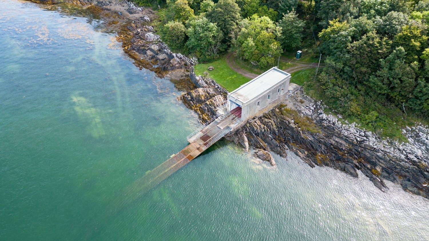 A rectangular concrete building with a long slipway on a rocky foreshore. There are lots of trees behind the station and the water is greeny-blue.