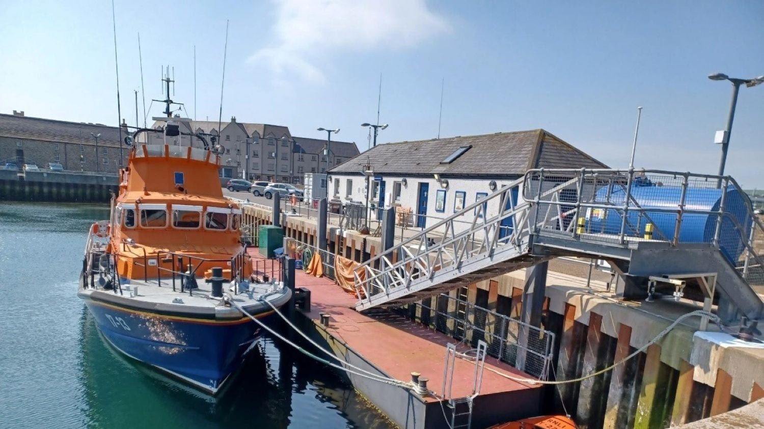 A one storey grey building with blue doors next to a pontoon where an all-weather lifeboat is moored.