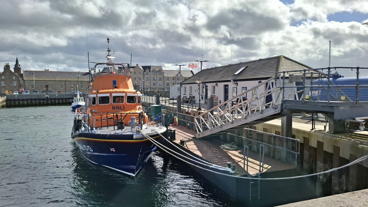 A one storey grey building with blue doors next to a pontoon where an all-weather lifeboat is moored.