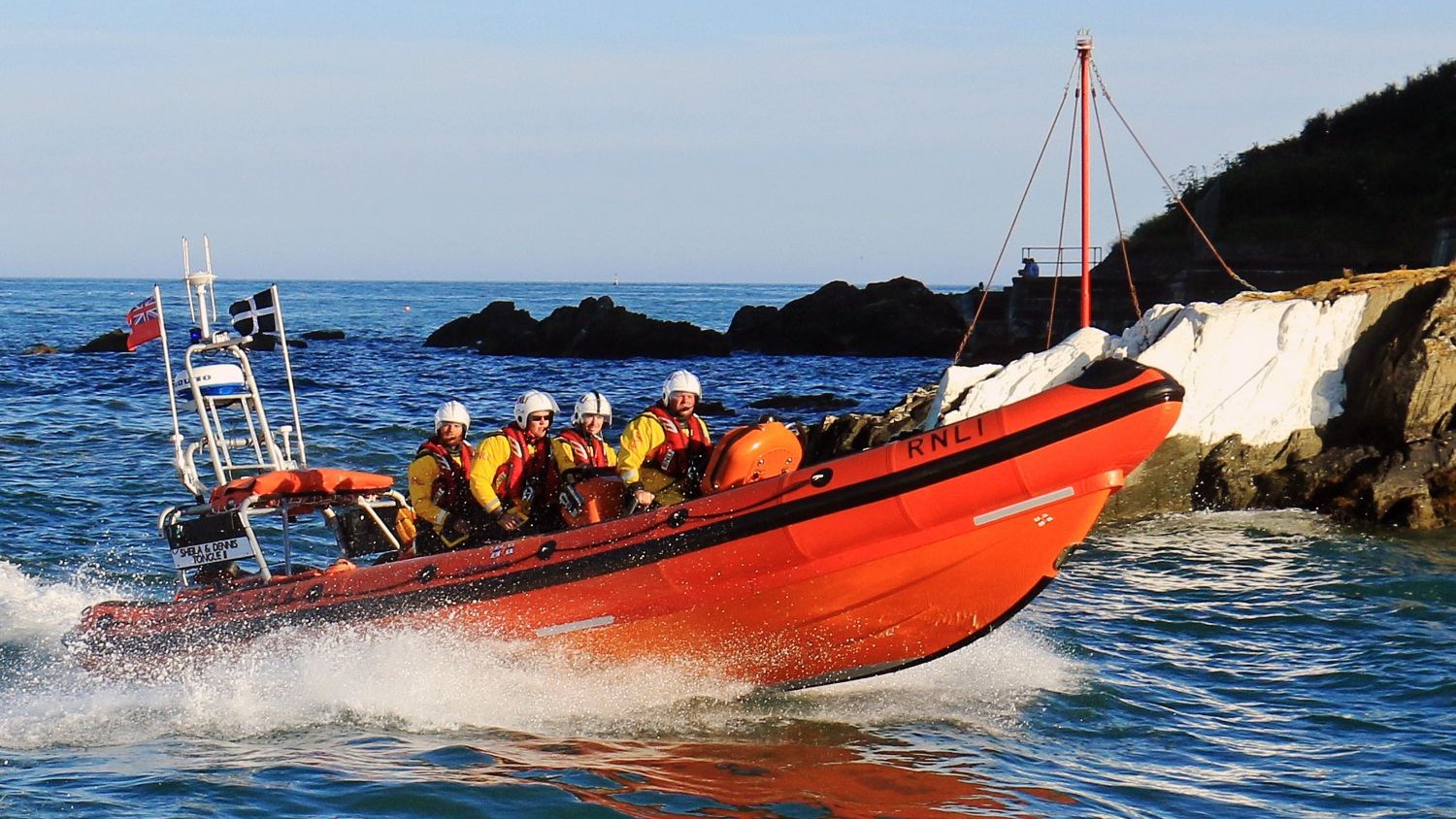 A large rib with four crew creating spray on calm blue waves.