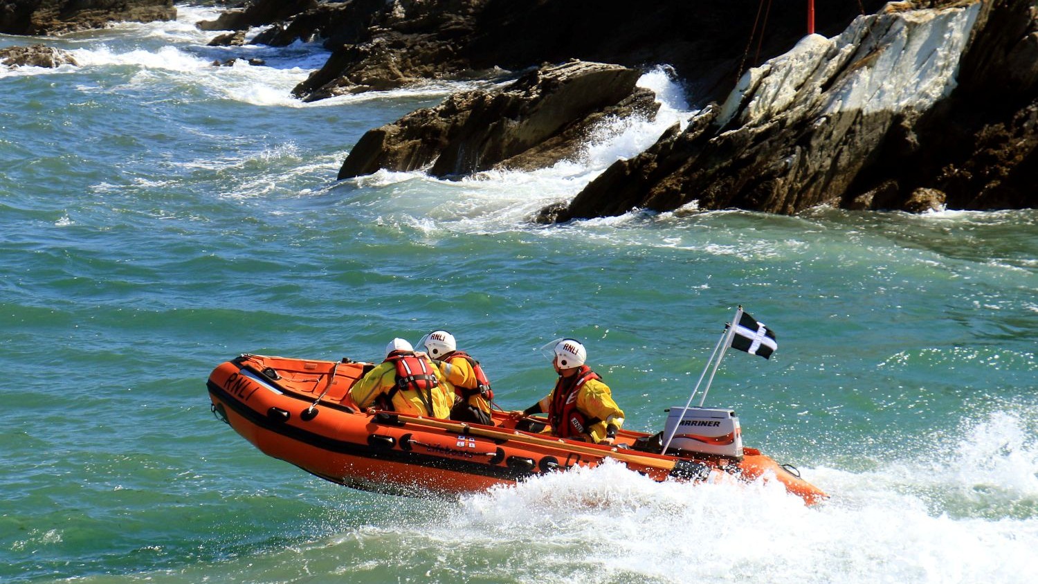 A small rib with three crew crests a green wave with a rocky outcrop behind them.