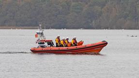 An RNLI lifeboat named "Jean Spier" with five crew members in yellow jackets and helmets travels across calm water, with trees and wildlife visible in the background.