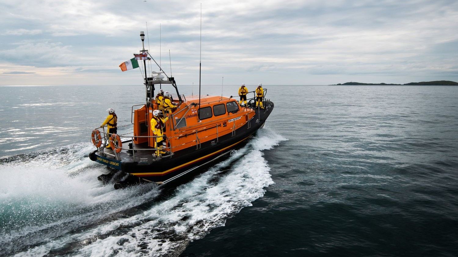 An orange RNLI lifeboat travels across calm water, leaving a curved white wake behind it. Several crew members in yellow jackets, red lifejackets, and white helmets stand on deck. The sky is overcast, and small islands or landmasses are visible on the horizon.