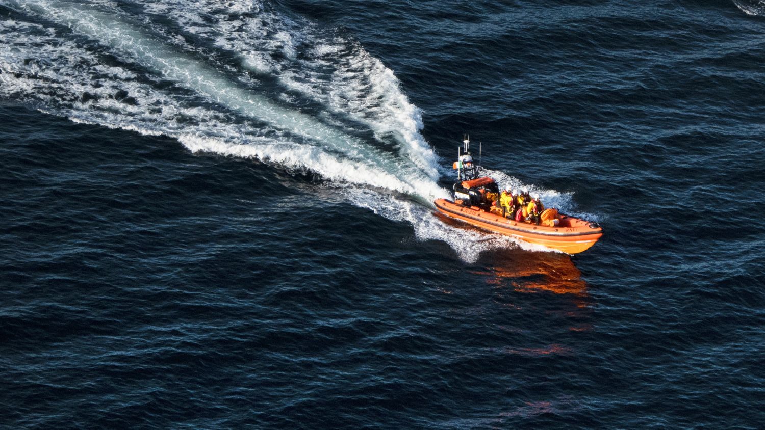 Aerial view of a lifeboat as it speeds across dark blue water, leaving a curved trail of white foamy wake behind.