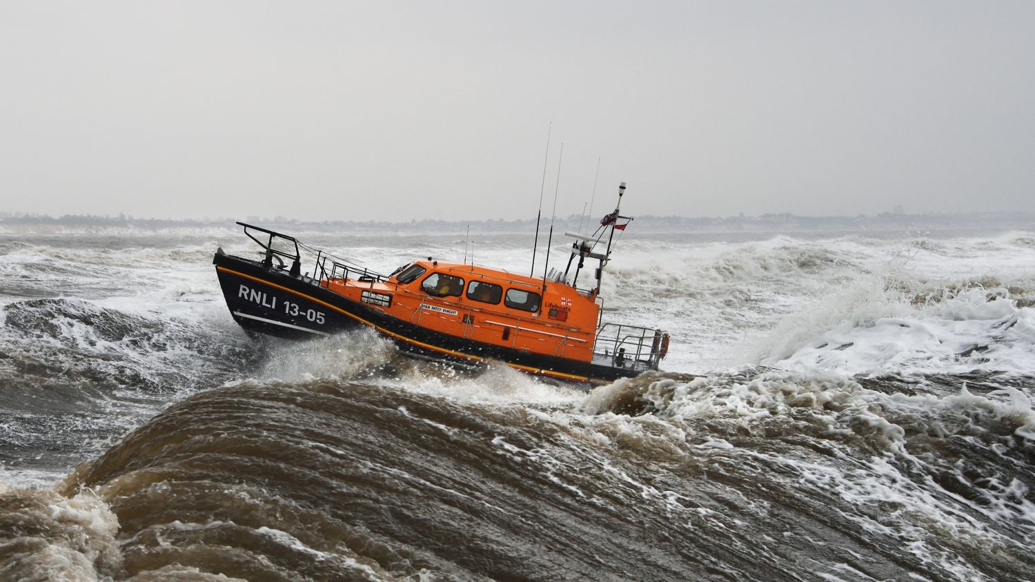 RNLI lifeboat 13-05 battling through large waves in rough grey seas, angled upward as it rides over a crest