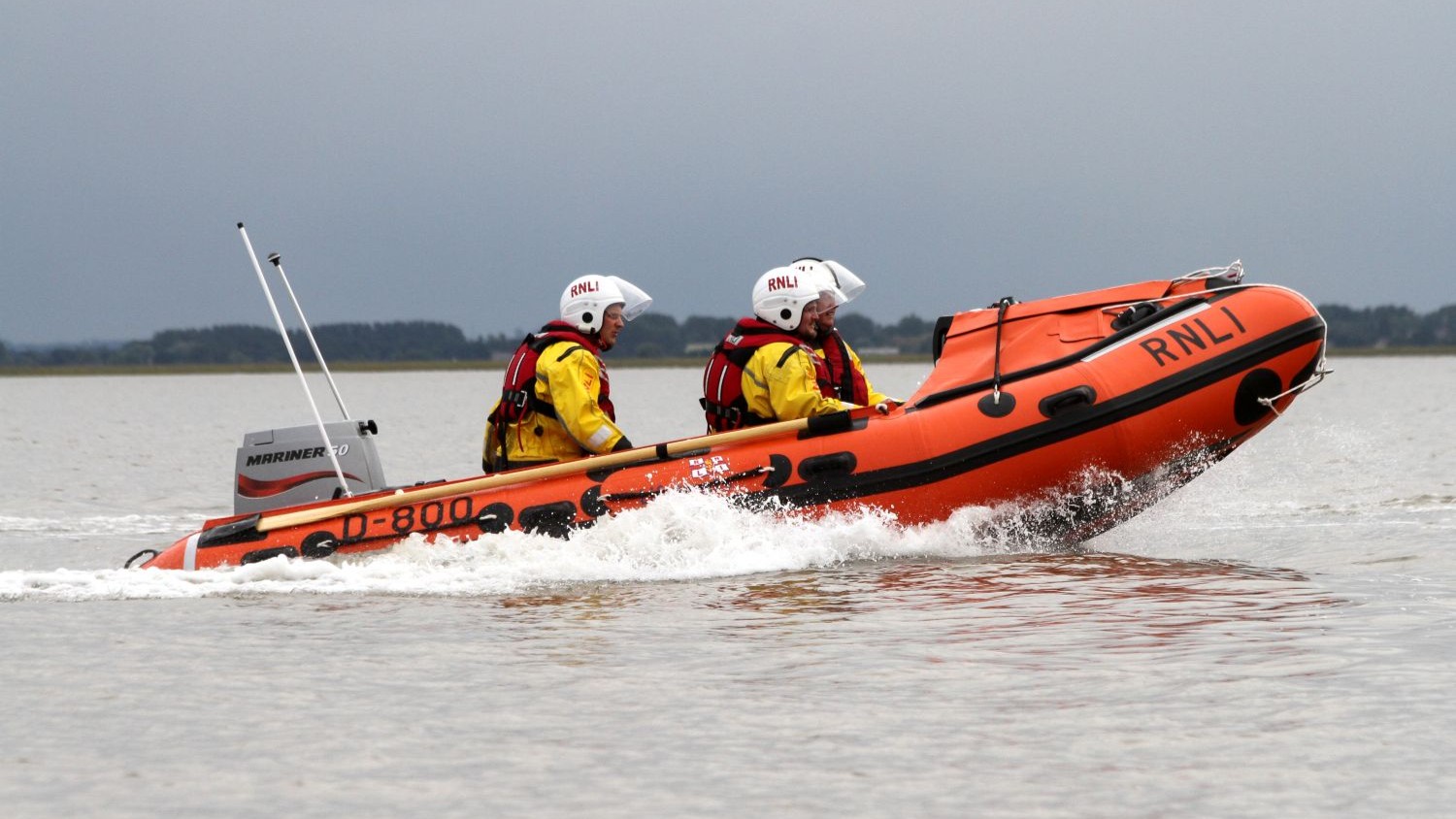 An orange RNLI inflatable lifeboat moves across shallow water from left to right, creating a small spray. Two crew members wearing yellow jackets, red lifejackets, and white helmets sit facing forward. The sky is overcast, and a low, distant shoreline is visible in the background.