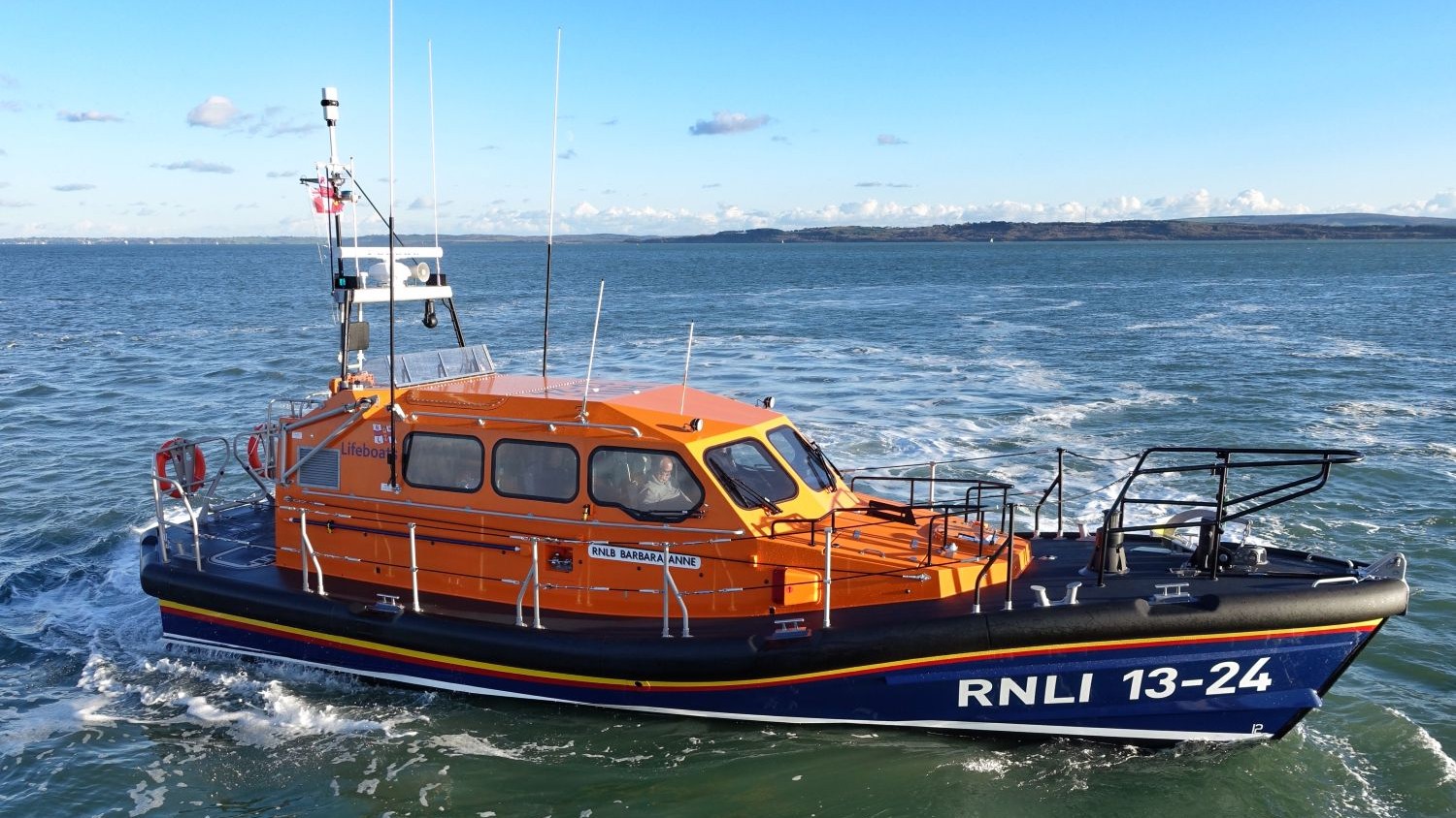 An orange RNLI lifeboat marked “RNLI 13-24” moves across calm blue water. The boat has a dark blue hull and multiple antennas on top. White spray trails behind it. A distant shoreline is visible under a clear sky with scattered clouds.