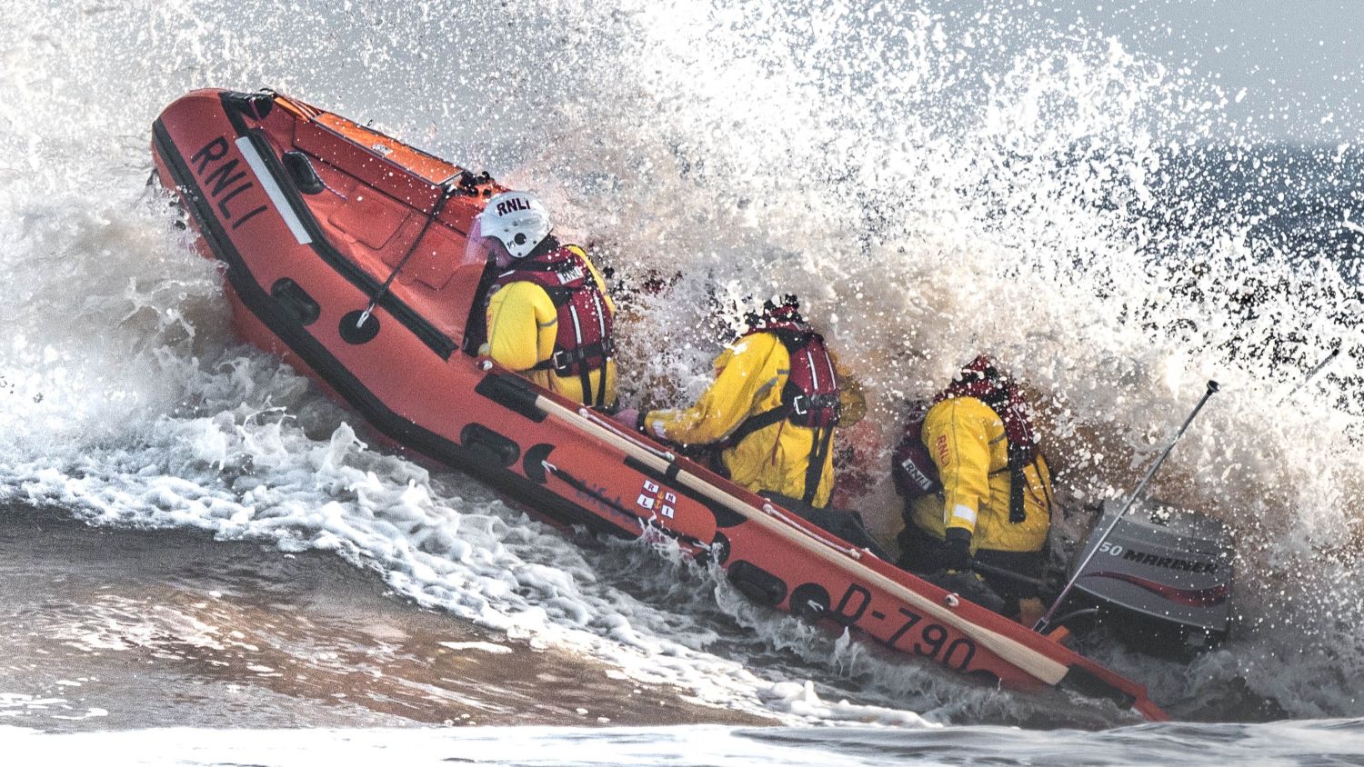 Orange RNLI lifeboat with three crew members in yellow gear powering through crashing waves near shore.