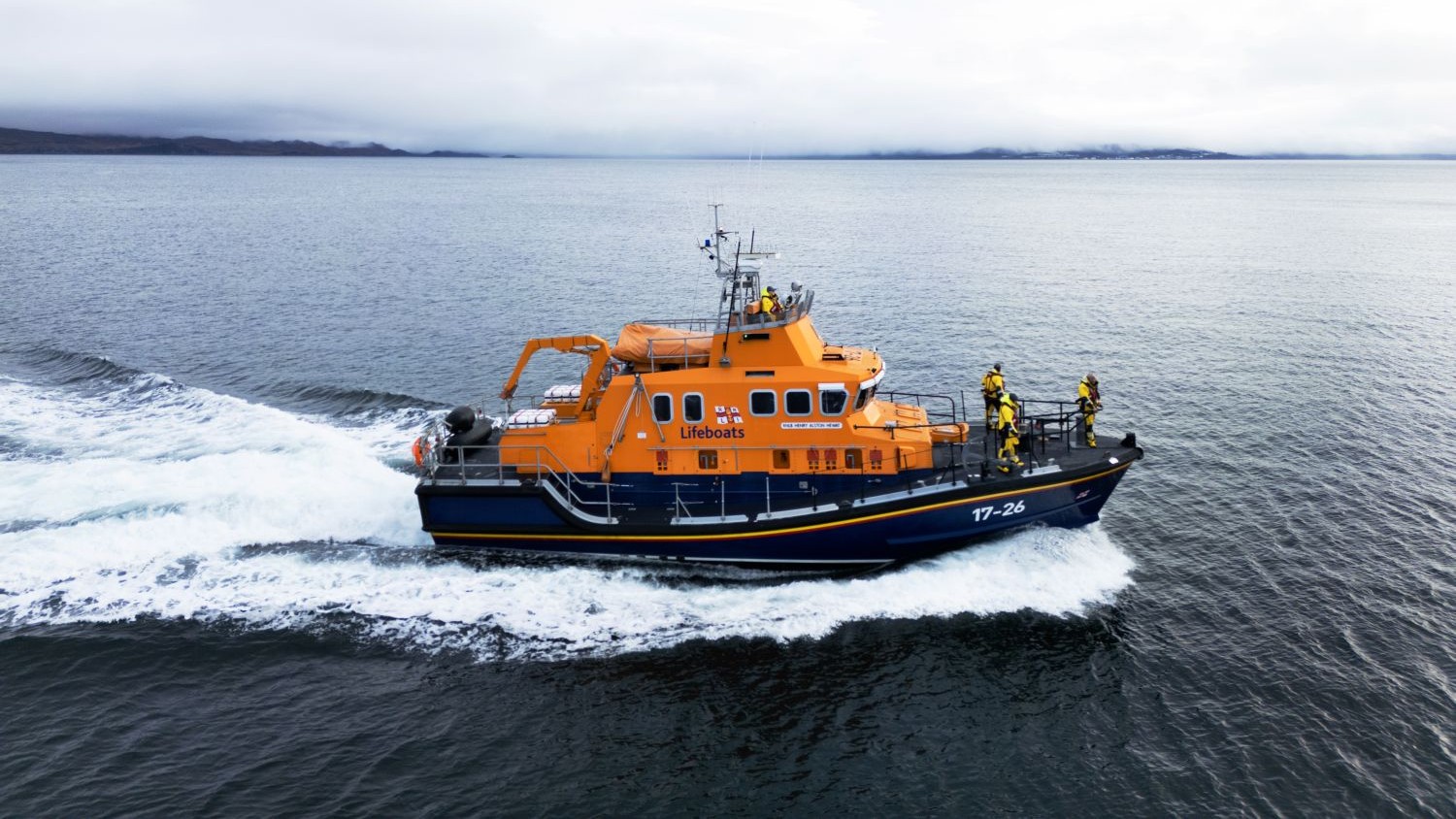 An orange RNLI lifeboat marked “17‑26” travels across calm water, leaving a white wake behind it. Several crew members in yellow jackets, red lifejackets, and white helmets stand on deck. The sky is overcast, and a low, distant shoreline is visible on the horizon.