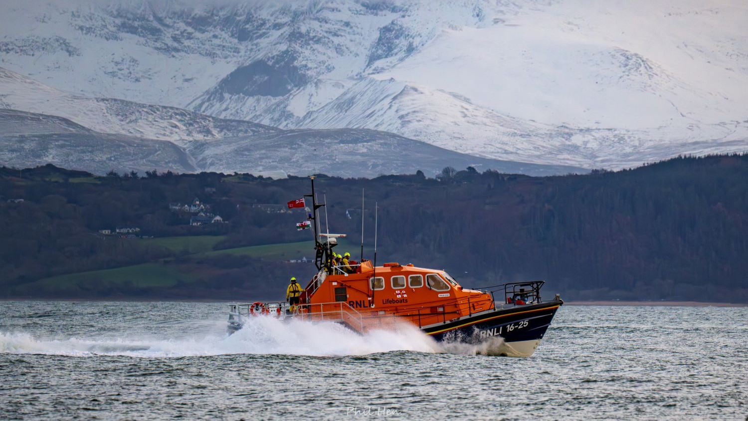 An orange RNLI lifeboat speeds across rough grey water, leaving a trail of white spray. Snow-covered mountains rise in the background behind the boat and distant shoreline.