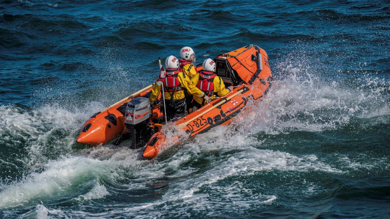 A small orange RNLI inflatable lifeboat rides over choppy dark-blue waves, sending up white spray. Three crew members in yellow jackets and white helmets face forward as the boat speeds through the water.