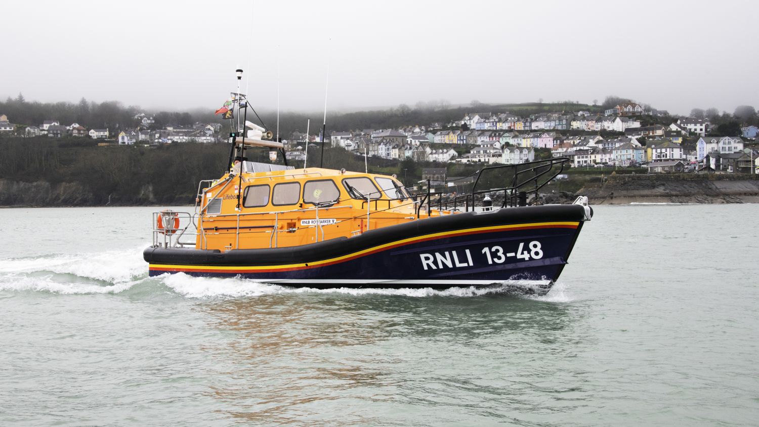 A lifeboat speeds across choppy water, creating white spray behind it. In the background, a coastal town with rows of white buildings lines a hillside under an overcast sky.