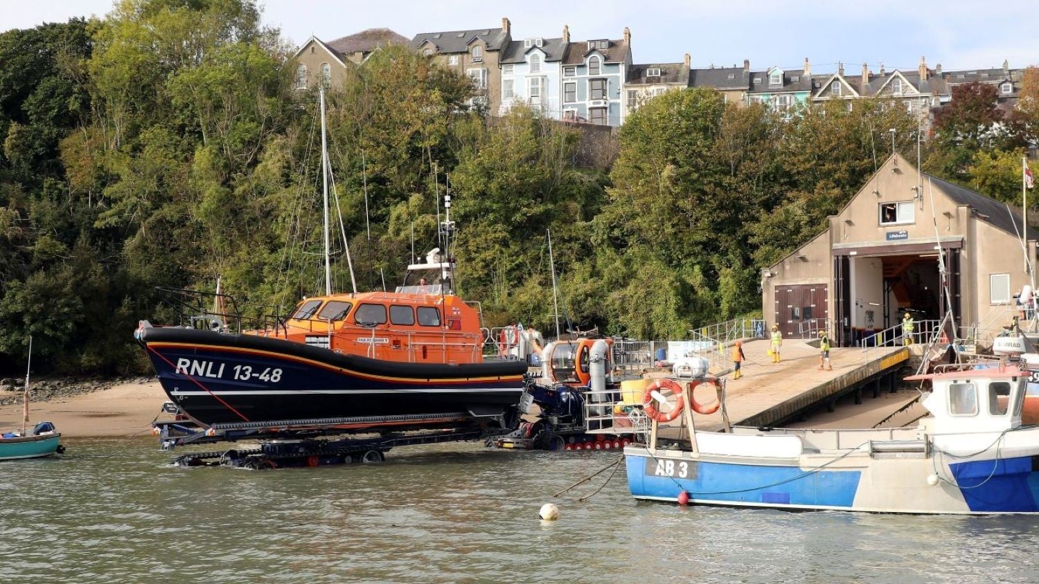 A cream building with pointy roof, the boathouse doors open onto a slipway where a tractor is pushing out a Shannon class lifeboat.
